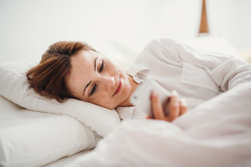 A young woman lying in bed indoors in the morning in a bedroom, using smartphone.