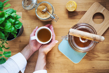 A composition of hands holding cup of tea, teapot and sugar on wooden table. A top view.