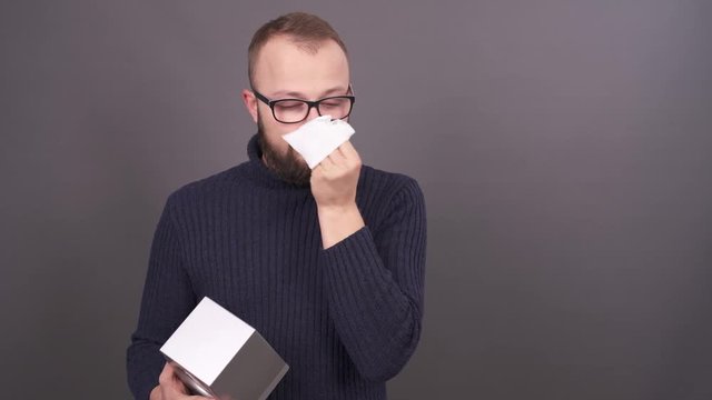 Close-up portrait of a bearded young snotty guy in glasses blowing his nose in napkins. Isolated on grey background.