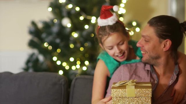 Cute schoolgirl in a rim with a Santa hat gives her father a Christmas gift in a gold box. Happy family christmas