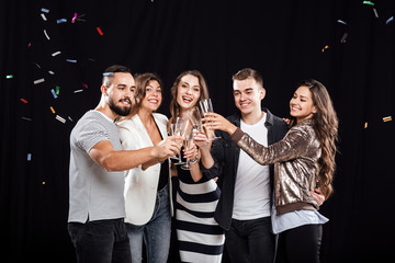 Company of friends in stylish casual clothes stand together and clink glasses with champagne on a black background and confiture around. Party time