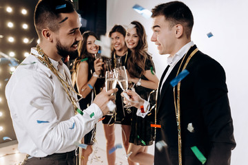 Beautiful young girls and guys dressed in stylish elegant clothes smile  together and clink glasses with champagne confiture around. Party time