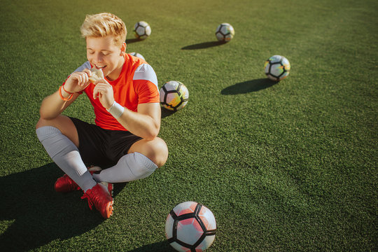 Happy young player sit on lawn with legs crossed and eat sandwich. He is alone. Four balls are behind hum and one is in front. Sunny weather outside. - Powered by Adobe