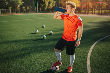 Picture of young football player stand on lawn and drink water. He hold feet on ball. Another four...