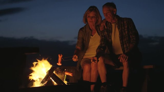Happy Caucasian American seniors on camping vacation cooking sages over a campfire flames outdoors 