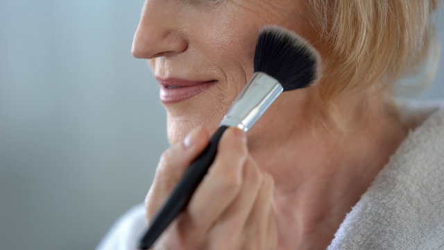 Mature woman applying face powder by make-up brush, preparing for date, beauty
