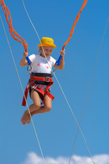A little girl jumps high on a trampoline with rubber ropes against the blue sky and white clouds. Adventures and extreme sports. Concept of summer vacation, jumping.