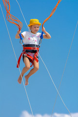 A little girl jumps high on a trampoline with rubber ropes against the blue sky and white clouds. Adventures and extreme sports. Concept of summer vacation, jumping.