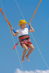A little girl jumps high on a trampoline with rubber ropes against the blue sky and white clouds. Adventures and extreme sports. Concept of summer vacation, jumping.