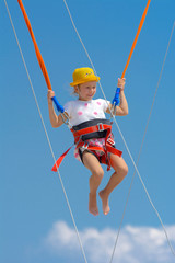 A little girl jumps high on a trampoline with rubber ropes against the blue sky and white clouds. Adventures and extreme sports. Concept of summer vacation, jumping.