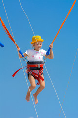 A little girl jumps high on a trampoline with rubber ropes against the blue sky and white clouds. Adventures and extreme sports. Concept of summer vacation, jumping.
