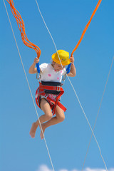 A little girl jumps high on a trampoline with rubber ropes against the blue sky and white clouds. Adventures and extreme sports. Concept of summer vacation, jumping.