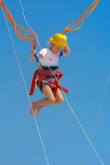 A little girl jumps high on a trampoline with rubber ropes against the blue sky and white clouds. Adventures and extreme sports. Concept of summer vacation, jumping.