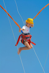 A little girl jumps high on a trampoline with rubber ropes against the blue sky and white clouds. Adventures and extreme sports. Concept of summer vacation, jumping.