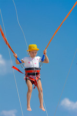 A little girl jumps high on a trampoline with rubber ropes against the blue sky and white clouds. Adventures and extreme sports. Concept of summer vacation, jumping.