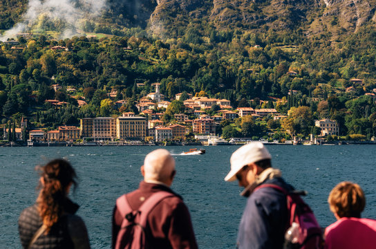 Tourists watch how boat moves against Cadenabbia town with buildings and hotels, coast of beautiful Como lake, Italy.