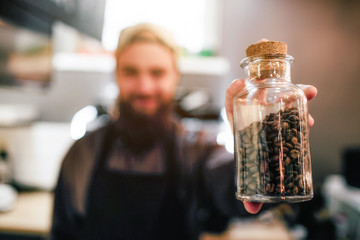 Happy young bearded barista hold glass jar with coffee beans. He shows it on camera. Young man stand in room.