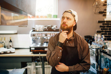 Young thoughtful man stand and think. He hold hand under chin. Guy is in kitchen. Light comes out of window.