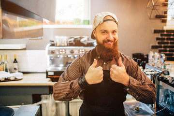 Happy young barista stand and show big thumb up on both hands. He look on camer. Guy stand in...