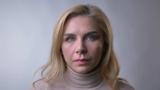 Close-up portrait of middle-aged blonde lady watching seriously into camera on gray background.