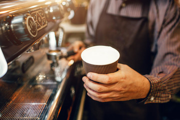 Cut view of barista in apron holds cup of coffee with floating milk. He stands at coffee machine.