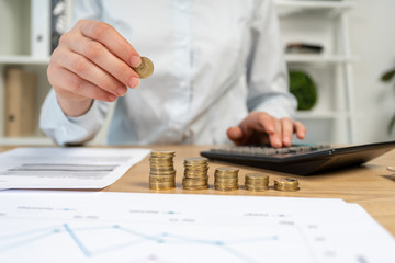 Close up cropped sale manager or marketer agent lady in her formal wear shirt she put coins on pile sit behind desk with charts graph graphics in light loft interior workplace use calculator