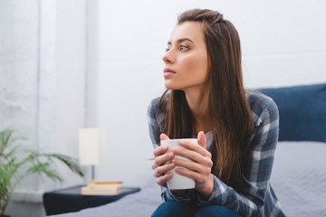pensive young woman holding cup and looking away while sitting at home