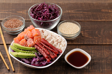 poke bowl Hawaiian food. a plate of rice, salmon, avocado, cabbage and cheese, next to sesame and soy sauce. on a brown wooden table.