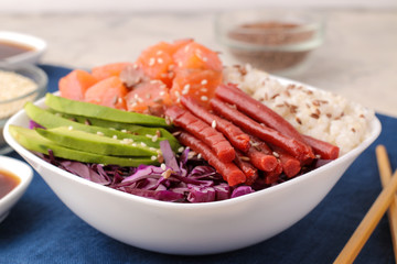 poke bowl Hawaiian food. a plate of rice, salmon, avocado, cabbage and cheese. near skunk and soy sauce on a blue napkin on a light background.