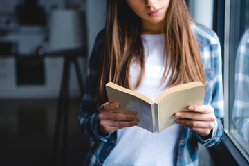 cropped shot of young woman standing near window and reading book at home