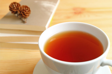 Closed up a cup of hot tea on wooden table with blurred stack of books and dry pine cones on top in background 