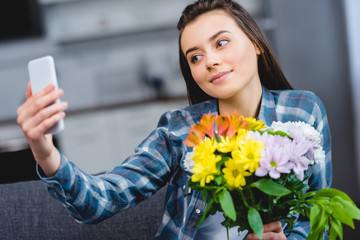 beautiful smiling young woman holding flowers and taking selfie with smartphone at home