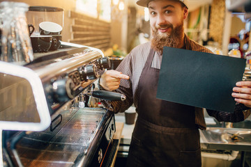 Excited young man points on piece of dark paper. He hold it in hand and look on camera. Guy stand at coffee machine in kitchen.