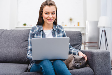 Naklejka premium beautiful smiling young woman using laptop and stroking cat at home