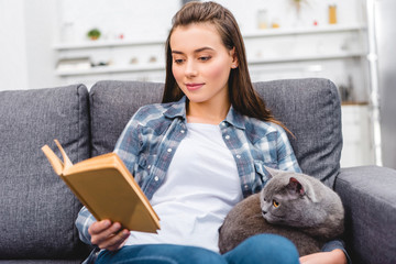 smiling young woman reading book and sitting with cat on couch
