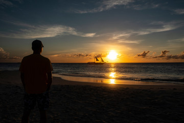 Silhouette of a Man Watching Sunset Over the Ocean with a Sailboat Passing the Horizon