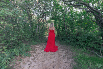 A young blonde woman in a red dress on enchanted woods going away or walking away
