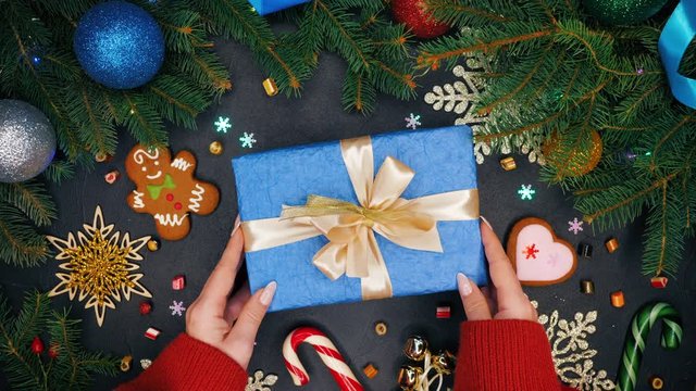 Hands of girl puts present wrapped in blue paper and then takes it. Black table with Christmas decorations - gingerbread, snowflakes, lollipops. New year festive mood