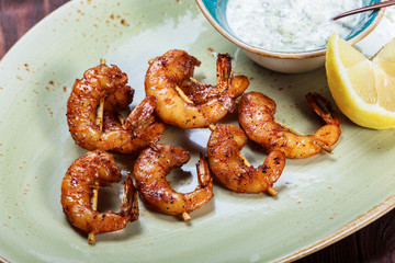 Roasted shrimps on skewers with sauce and lemon on plate on wooden table. Delicious dish of seafood