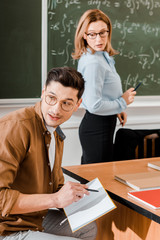 Young student looking away and holding notebook with pen near female teacher