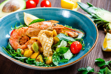 Warm salad with seafood, langoustine, mussels, shrimps, squid, scallops, mango, pineapple, avocado, arugula and basil on wooden background close up. Mediterranean food. Top view