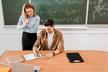 Student in glasses wtiting in notebook near teacher in classroom