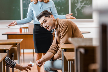 selective focus of dissatisfied female teacher looking at male student taking crib during exam in...