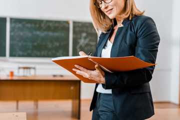 cropped view of female teacher in formal wear writing in notebook in classroom