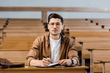 focused male student in glasses sitting at desk, looking at camera while writing in notebook during lesson in classroom