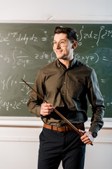 smiling male teacher in formal wear holding wooden pointer in front of chalkboard with equations