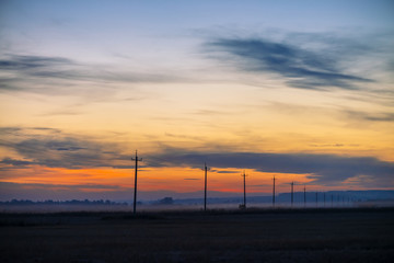 Fototapeta premium Power lines in field on sunrise background. Silhouettes of poles with wires at dawn. Cables of high voltage on warm orange blue sky. Power industry at sunset. Multicolored picturesque vivid sky.