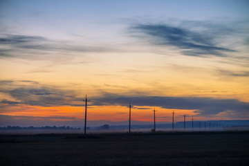 Power lines in field on sunrise background. Silhouettes of poles with wires at dawn. Cables of high voltage on warm orange blue sky. Power industry at sunset. Multicolored picturesque vivid sky.