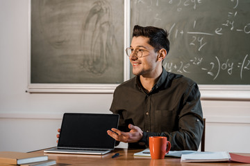 smiling male teacher sitting at desk and showing laptop with blank screen in classroom