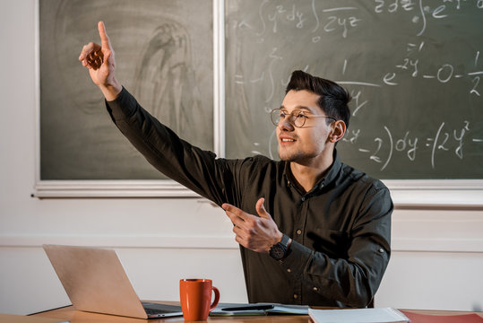 male teacher sitting at desk, pointing finger in air and explaining equations in maths classroom